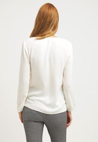 Weiße Bluse mit langen Ärmeln aus glattem, leichtem Stoff. Verfügt über einen abgerundeten Saum und ein dezentes Rückennaht-Detail. Kombiniert mit grauen Hosen.