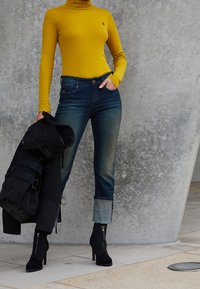 Gelbes langärmliges Turtleneck, blaue Jeans mit hochgerollten Säumen, schwarze Absatzstiefeletten, kombiniert mit einer schwarzen Jacke, die in einer Hand gehalten wird.