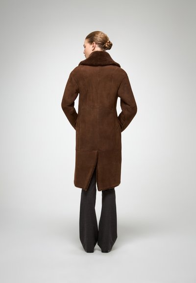 Brūns zamšādas mētelis ar lielu mākslīgā kažoka apkakli, vidēja garuma dizains, sānu kabatas un aizmugures atvērums mobilitātei. Tekstūrēts apdare.