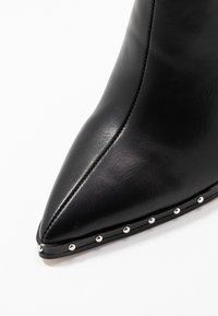 Musta nahkasaapas nilkkapituisena, teräväkärkinen, jossa hopeiset niitit kengän pohjassa. Sileä pinta ja virtaviivainen muotoilu.