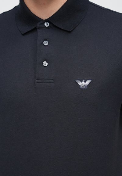 Čierne polo tričko s textúrovanou tkaninou, vybavené zapínaním na tri gombíky a malým vyšívaným logom na ľavej strane hrudníka.