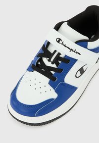 Μπλε και λευκό αθλητικό παπούτσι Champion με μαύρα κορδόνια και λωρίδα βέλκρο σε λευκό φόντο, που παρουσιάζει λεπτομέρειες από μπροστά και πλάγια.