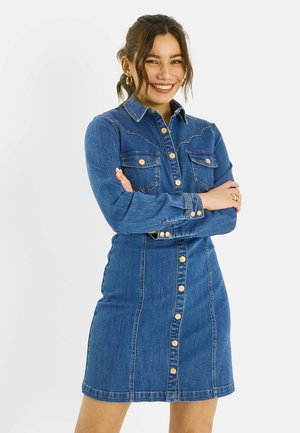 Mujer sonriendo con los brazos cruzados, vistiendo un vestido vaquero abotonado con mangas largas y botones dorados, sobre un fondo blanco liso.