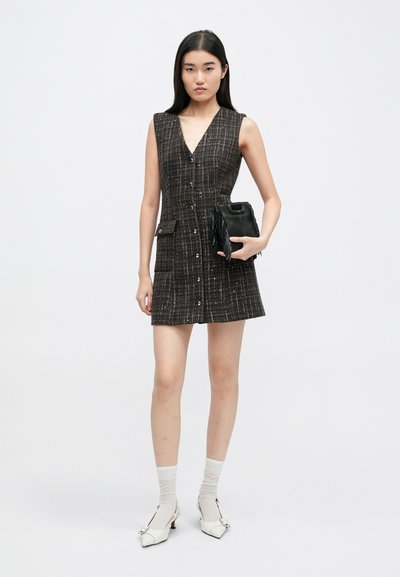 Vestido sin mangas a cuadros en blanco y negro con cierre de botones y dos bolsillos laterales. La modelo sostiene un bolso negro con flecos, vistiendo calcetines blancos y tacones.