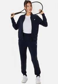 Ciemnogranatowy sportowy dres wykonany z miękkiej tkaniny, z kurtką na zamek i dopasowanymi spodniami. Pod spodem biała koszulka i białe trampki.