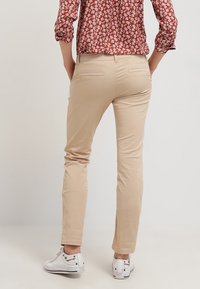 En person iført beige slim-fit bukser, en rød blomstret langærmet skjorte og hvide sneakers, står med hænderne i baglommen.