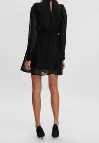 Robe noire transparente à manches longues, avec des accents à volants, un détail de dos en forme de trou de serrure, et une taille cintrée, associée à des chaussures noires à talons hauts.