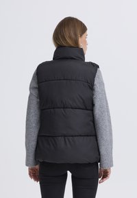 Opgeblazen zwart vest met een hoge kraag, doorgestikt ontwerp en mouwloze snit, gecombineerd met een grijze trui met lange mouwen en donkere spijkerbroek.