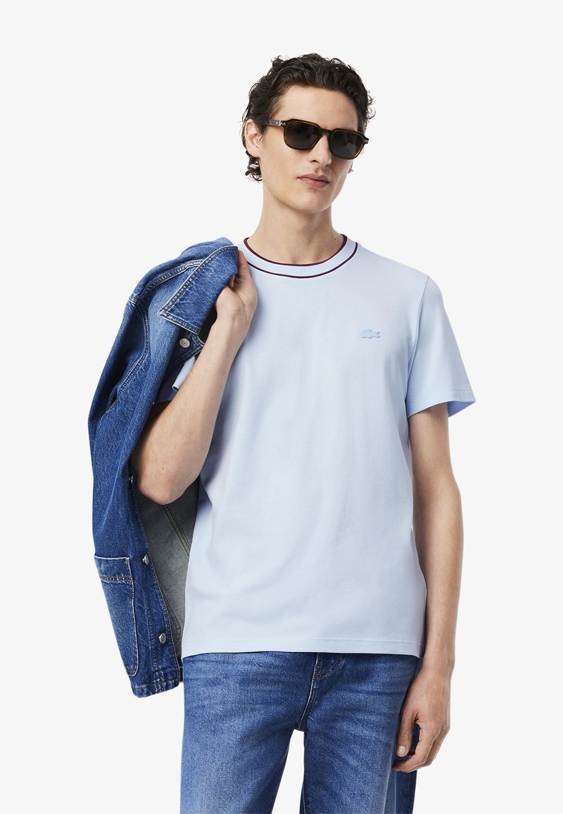 T-shirt in cotone azzurro chiaro con scollo rotondo e rifiniture bordeaux. Indossato con una giacca di jeans, che mostra uno stile casual a strati.