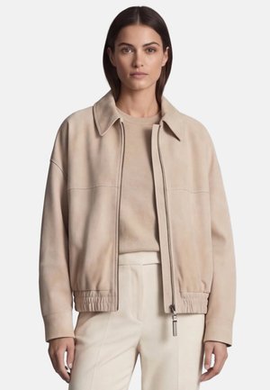 Vrouw draagt een beige jas met rits over een bijpassende trui en lichte broek, staand tegen een effen achtergrond.