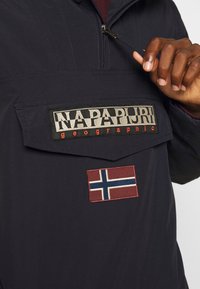 Osoba ubrana w ciemną kurtkę z suwakiem z przodu, trzymająca za suwak, z naszywką logo Napapijri oraz naszywką norweskiej flagi.