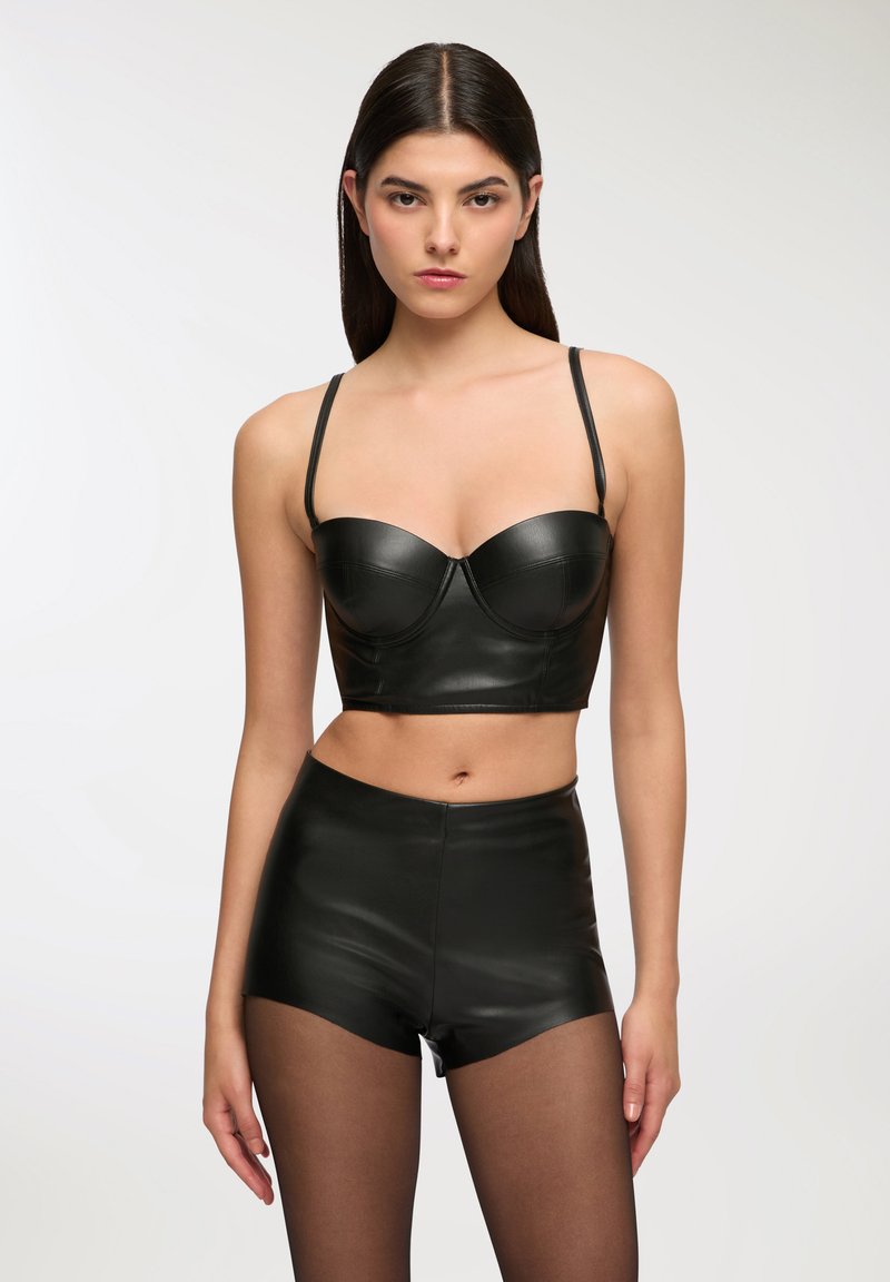 Top bustier corto in pelle nera con spalline sottili e coppe strutturate, abbinato a pantaloni corti in pelle nera a vita alta e collant neri trasparenti.