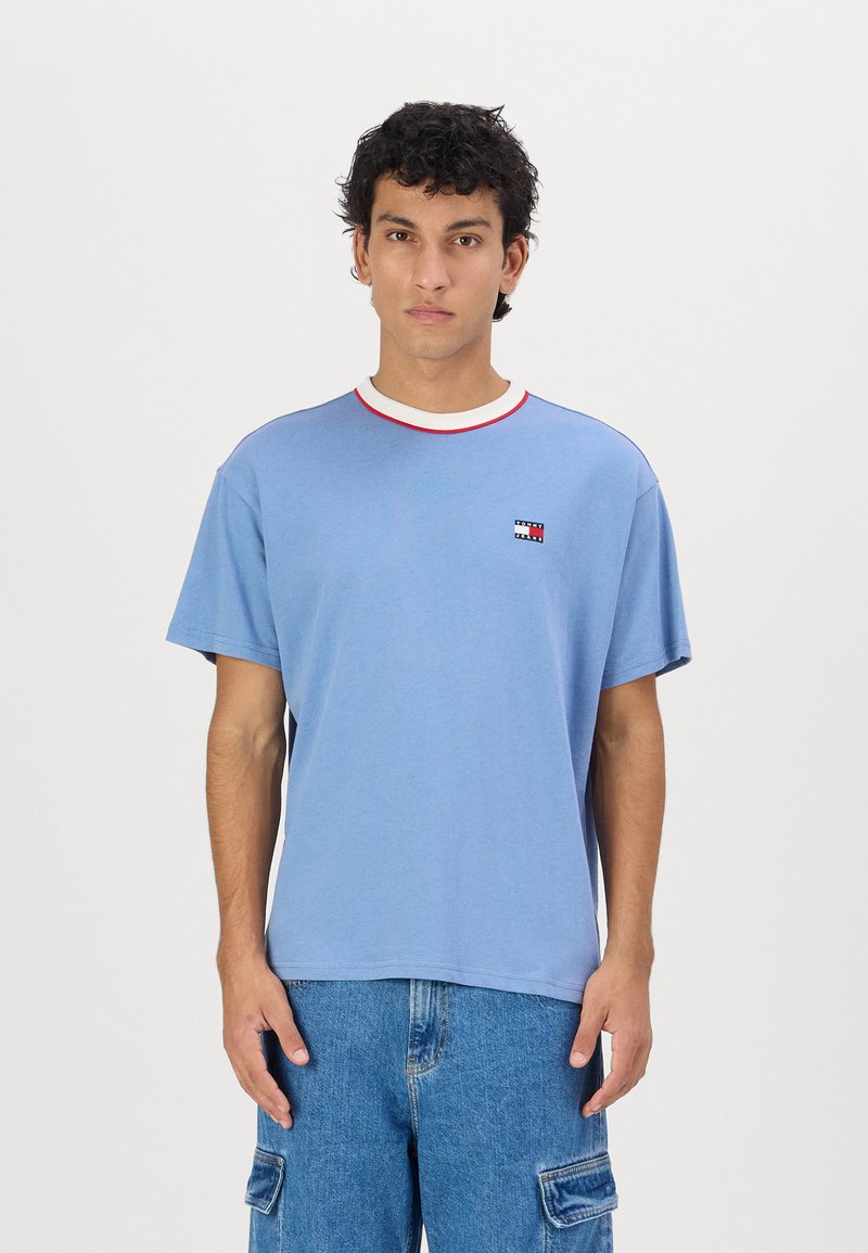Camiseta de algodón azul claro con mangas cortas, cuello acanalado blanco y rojo en contraste, que presenta un pequeño logo en el lado izquierdo del pecho.