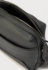 Fekete texturált bőrtok, részben nyitott ezüst cipzárral és apró bőr húzófüllel, világos háttéren.