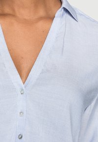 Lyseblå skjorte med krave og V-hals, der har en glat tekstur, fin syning og små, runde knapper langs fronten.