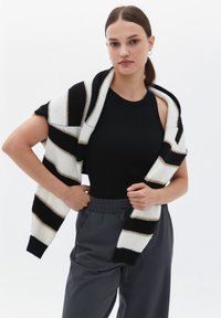 Cardigan de punto con gruesas franjas en blanco y negro, que presenta un corte relajado y un delantero abierto. El modelo lo combina con una blusa negra y pantalones grises.