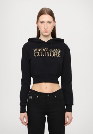 Schwarze, verkürzte Kapuzenjacke mit Kordelzug, versehen mit goldfarbenem Pailletten-Text "VERSACE JEANS COUTURE." Kombiniert mit schwarzen Jeans, die eine glatte Textur aufweisen.