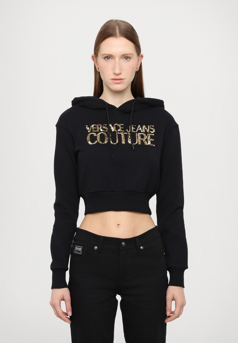 Zwarte cropped hoodie met trekkoord, voorzien van gouden sequins tekst "VERSACE JEANS COUTURE." Gematcht met zwarte jeans, die een gladde textuur tonen.