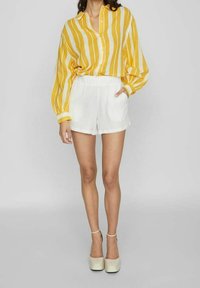 Žlutá a bílá pruhovaná košile s dlouhým rukávem a knoflíkovým zapínáním, v kombinaci s bílými šortkami. Modelka má na sobě béžové platformové boty.