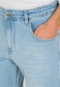 Jeans de mezclilla azul claro con una pretina estándar, botón metálico y bolsillos delanteros inclinados. Tela texturizada con detalles de costura.