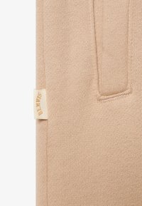 Beige strikket stoff har en lomme og en vevd etikett med teksten "Skavis" i lys gull. Teksturen er myk og litt elastisk.
