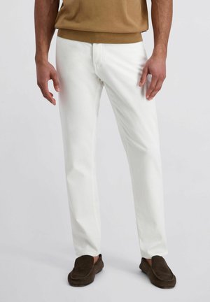 Mann trägt weiße, gerade geschnittene Hosen, braune Slipper und einen beige Pullover, steht mit entspannt an den Seiten herunterhängenden Händen.