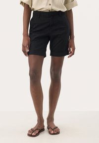 Svarta bomullsshorts med uppvikta fållar, med framfickor och en åtsittande midja. Matchat med bruna flip-flops på en ljus yta.