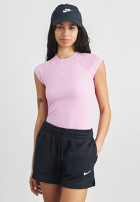 Egy nő rózsaszín ujjatlan Nike fels�őt, fekete Nike rövidnadrágot és fekete Nike sapkát visel, egyik kezét zsebében tartva, egyszínű háttér előtt áll.