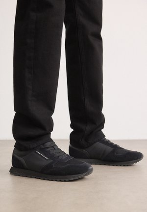 Personne portant des baskets noires décontractées et un pantalon noir, debout sur un sol lisse avec un fond clair uni.