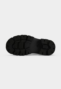 Semelle de chaussure en caoutchouc noir avec des crampons profonds et larges et des motifs texturés pour l’adhérence et la durabilité, présentée sur un fond blanc uni.