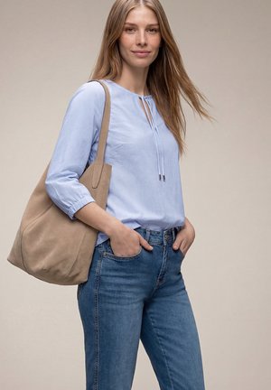 Kobieta w jasnoniebieskiej bluzce i dżinsach, z beżową torebką na ramieniu, ręce w kieszeniach, delikatnie uśmiechająca się na jednolitym tle.