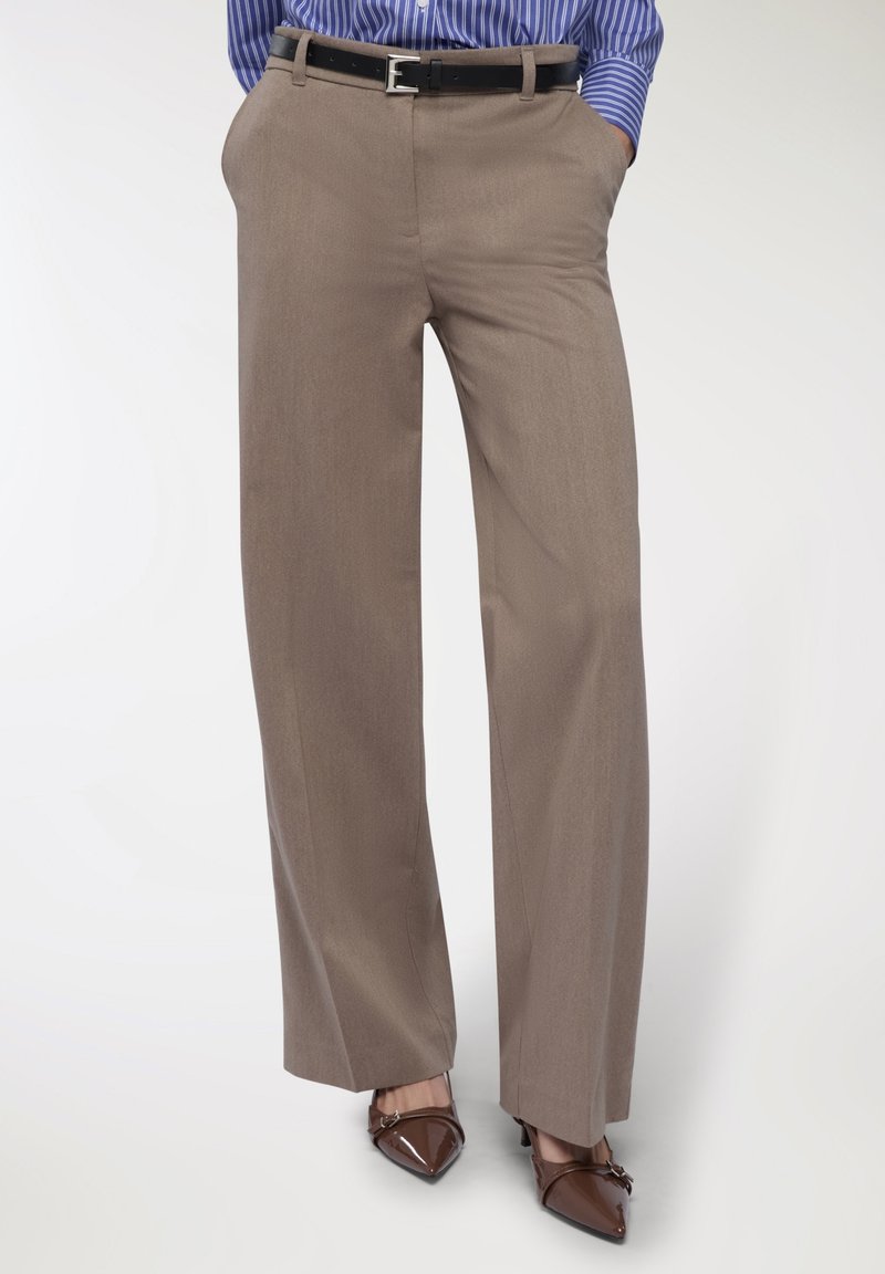 Osoba w beżowych szerokich spodniach z czarnym paskiem, ręce w kieszeniach, połączonych z brązowymi czubkowymi szpilkami i niebieską koszulą w paski.