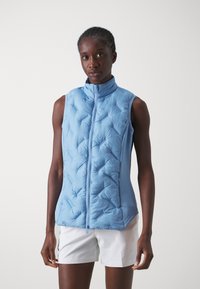 Žena u svijetloplavom prsluku bez rukava, zatvorenog zatvarača, u kombinaciji s bijelim kratkim hlačama, stoji ispred obične bijele pozadine.