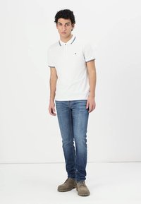 White polo shirt with navy trim detail, paired with blue jeans and beige shoes. The shirt features a small logo on the chest.