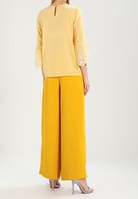 Gul rutig blus med utsvängda ärmar med spetskant, parat med livfulla gula vida byxor och blå högklackade sandaler.