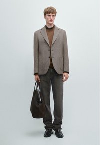 Brązowa marynarka w jodełkę, brązowy sweter, szare dżinsy, czarne buty, trzyma ciemnobrązową torbę typu tote. Prosty design z funkcjonalnymi kieszeniami.