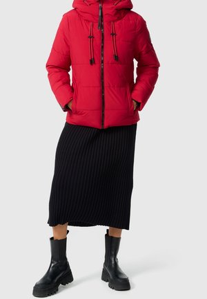 Osoba ubrana w czerwoną puchową kurtkę, czarną prążkowaną midi spódnicę oraz czarne botki na grubym obcasie, stojąca z rękami w kieszeniach.