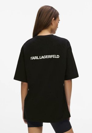 Kvinde med langt hår iført en løs sort T-shirt med "KARL LAGERFELD" printet i hvidt på ryggen mod en hvid baggrund.
