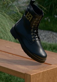 Schwarze Leder-Stiefelette mit runder Zehenpartie, goldenen Hardware-Akzenten und runden Schnürlöchern; verfügt über ein Logo am Schaft und eine dicke Sohle.