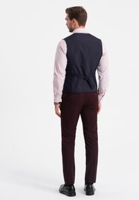 Gilet navy con una texture liscia, cinturino posteriore regolabile, indossato sopra una camicia rosa chiaro, abbinato a pantaloni bordeaux scuri e scarpe nere.