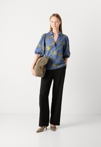 Modra cvetlična bluzica z nabranimi rokavi, v kombinaciji s črnimi širokimi hlačami. Model drži srednje veliko olivno zeleno torbico in nosi zlate škornje na peto.