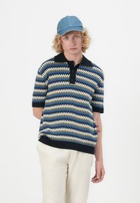 Polo à manches courtes avec un motif en zigzag aux couleurs bleu, vert et blanc. Col et ourlet noirs. Associé à un pantalon clair et une casquette en denim.