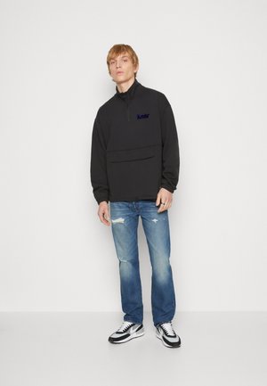 Pull en polaire noir avec col montant et demi-fermeture éclair, doté d'une poche frontale. Porté avec des jeans bleus déchirés et des baskets noires et blanches.
