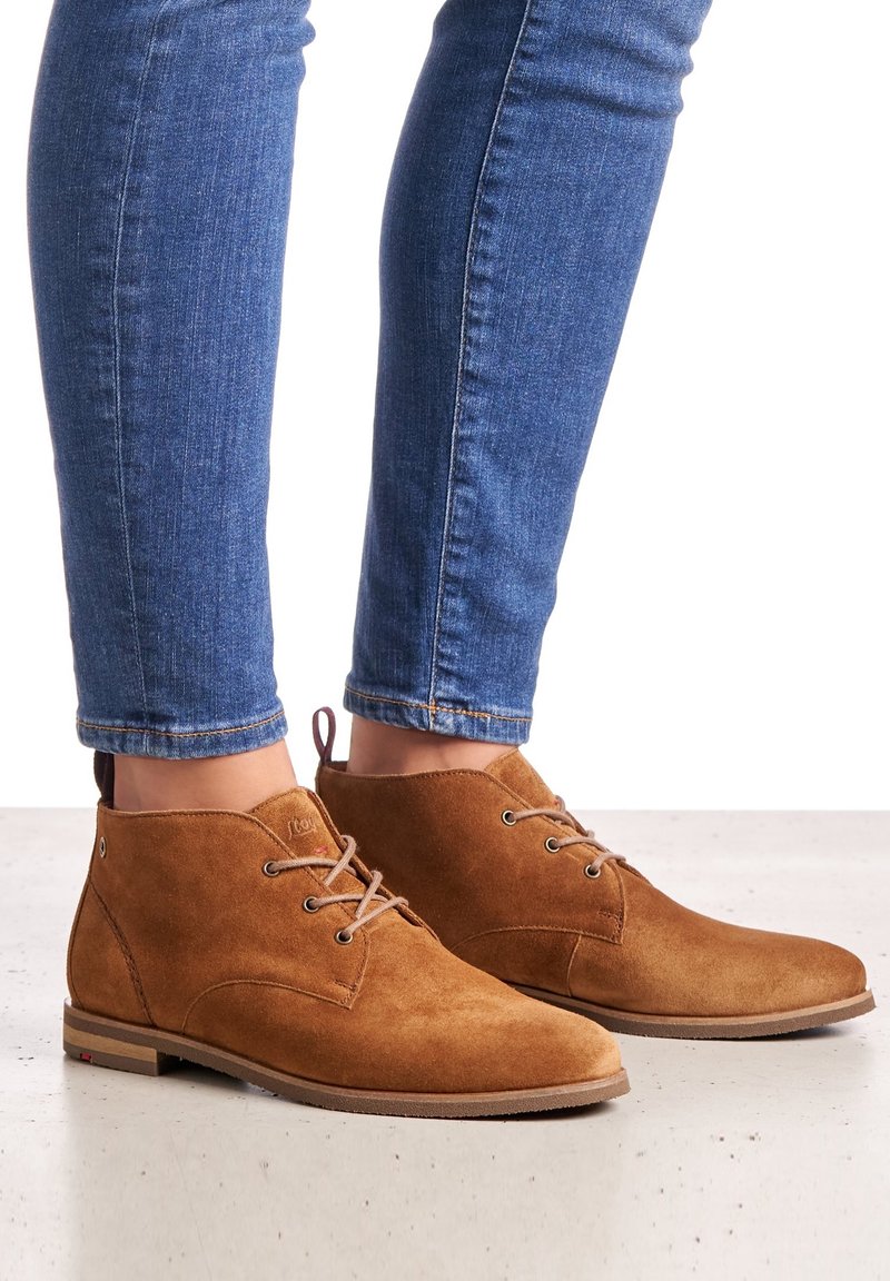 Botines de ante color marrón claro con una textura suave, puntera redonda y cordones marrón claro, combinados con jeans ajustados azules. Detalle de la suela de goma visible.