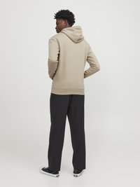 Beige hoodie with a front pocket and drawstring hood, paired with black trousers and black sneakers. The fabric has a smooth texture.