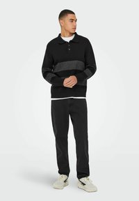 Schwarzer Strickpullover mit einem grauen horizontalen Streifen, versehen mit einem Kragen und einer dreiknöpfigen Knopfleiste. Kombiniert mit dunklen Hosen und hellen Sneakers.