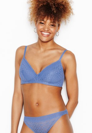Femme souriante aux cheveux bouclés portant un soutien-gorge et une culotte en dentelle bleue assortis, posant devant un fond blanc uni.