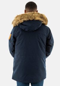 Giacca blu navy con cappuccio foderato in pelliccia sintetica, dotata di una toppa sulla manica sinistra e di un taglio più lungo con orlo dritto sul retro.