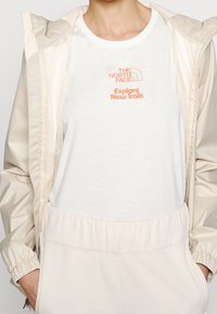 Maglietta leggera bianca con logo arancione ricamato "THE NORTH FACE" e "Explore New Trails" abbinata a una giacca beige e pantaloni.