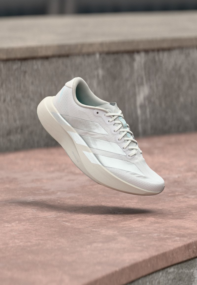 Chaussure de sport blanche à trois bandes suspendue en l'air au-dessus d'une surface en béton rosâtre et d'un fond en pierre grise.